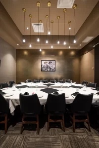 Front angle of an organized meeting room with a U-shaped table, tableware arranged on a white tablecloth, and contemporary decor in neutral and gold tones.