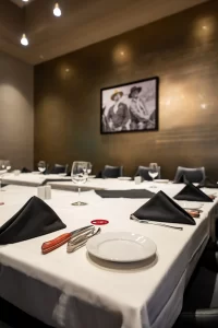 Detail of a table setting for events, focusing on white plates, black fabric napkins, cutlery, and glasses arranged on a white tablecloth.