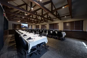 Wide shot of a spacious banquet room with a high vaulted ceiling, exposed wooden beams, and a long U-shaped table arrangement prepared for a formal event.