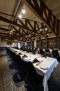 View of a long, narrow banquet room configured with a central table setup, highlighting the wood-trimmed windows and the warm, inviting lighting throughout the space.