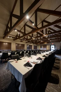 Wide-angle interior shot of a formal event hall with a high vaulted ceiling and exposed wooden beams, featuring a long U-shaped table setup with white tablecloths and black chairs.