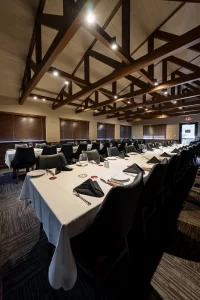 A long dining room set for a large group event, showcasing a long table arrangement under an impressive exposed wooden beam ceiling, with warm lighting and comfortable black chairs.