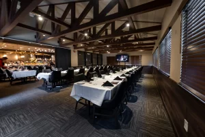 A banquet room set for a large meeting, focusing on the long rows of tables, black leather chairs, and the rustic charm of the exposed wooden beam ceiling.