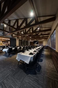 Wide view of an event hall prepared for a function, showcasing the long banquet table layout, floor-to-ceiling windows with wooden blinds, and an open entryway to the dining area.