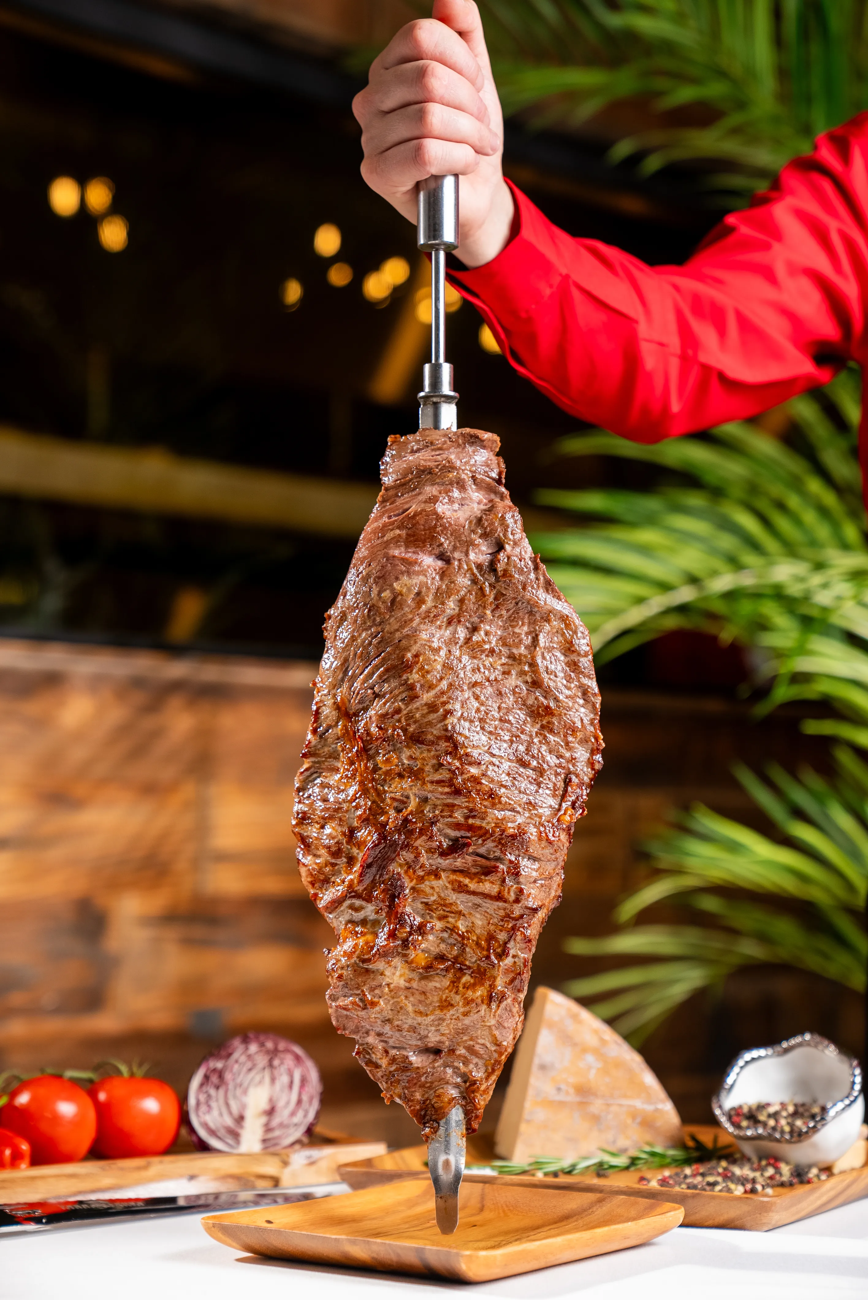 Large cut of grilled meat on a skewer held by a server in a red shirt.
