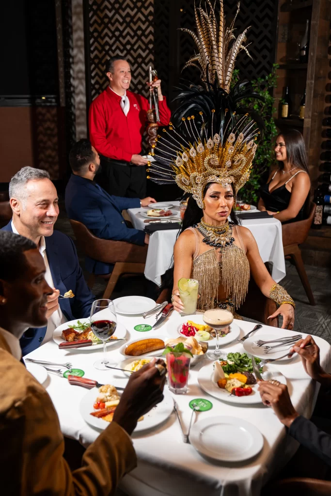 A lively dining scene with a woman in elaborate Carnival attire seated at a table, surrounded by guests enjoying a meal, while a server in red holds a skewer of meat.