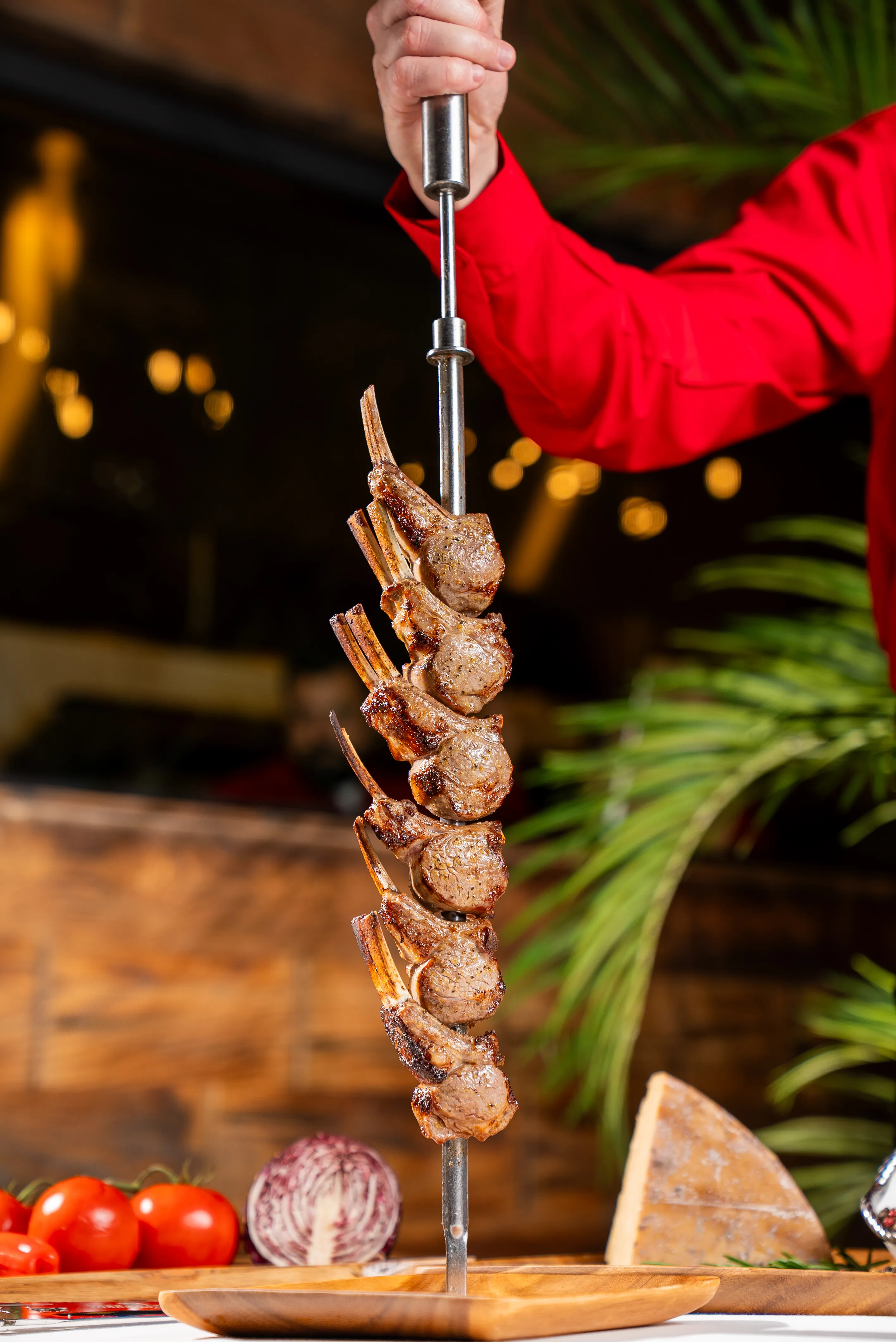 Skewered grilled lamb chops, each with a bone, held by a server in a red shirt.