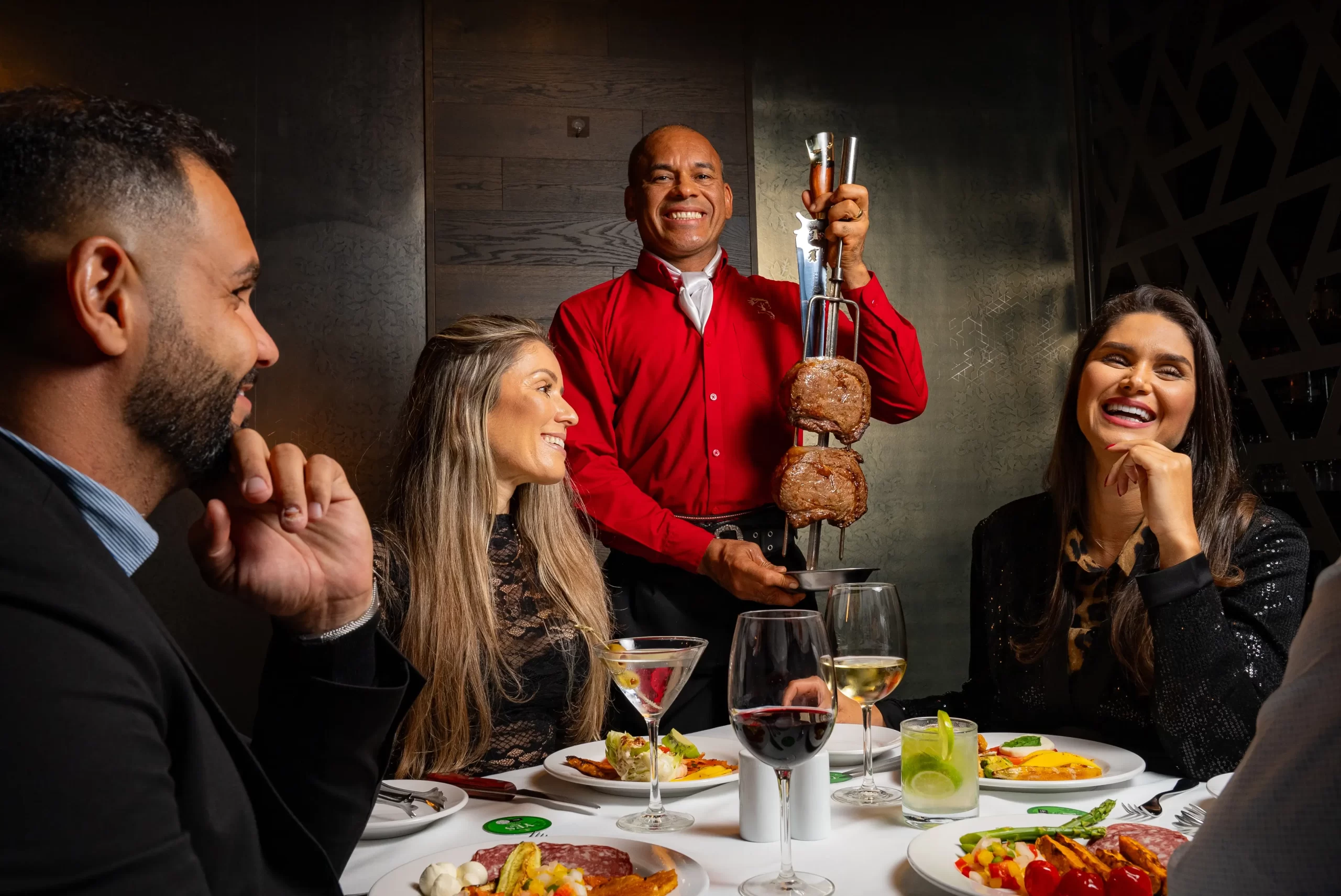 A group of people smiling as a server in a red shirt presents skewered grilled meat at their table.