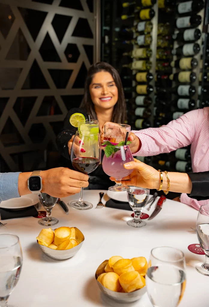 Three people clinking colorful drinks in celebration at a table, with small bowls of snacks and a wine rack in the background