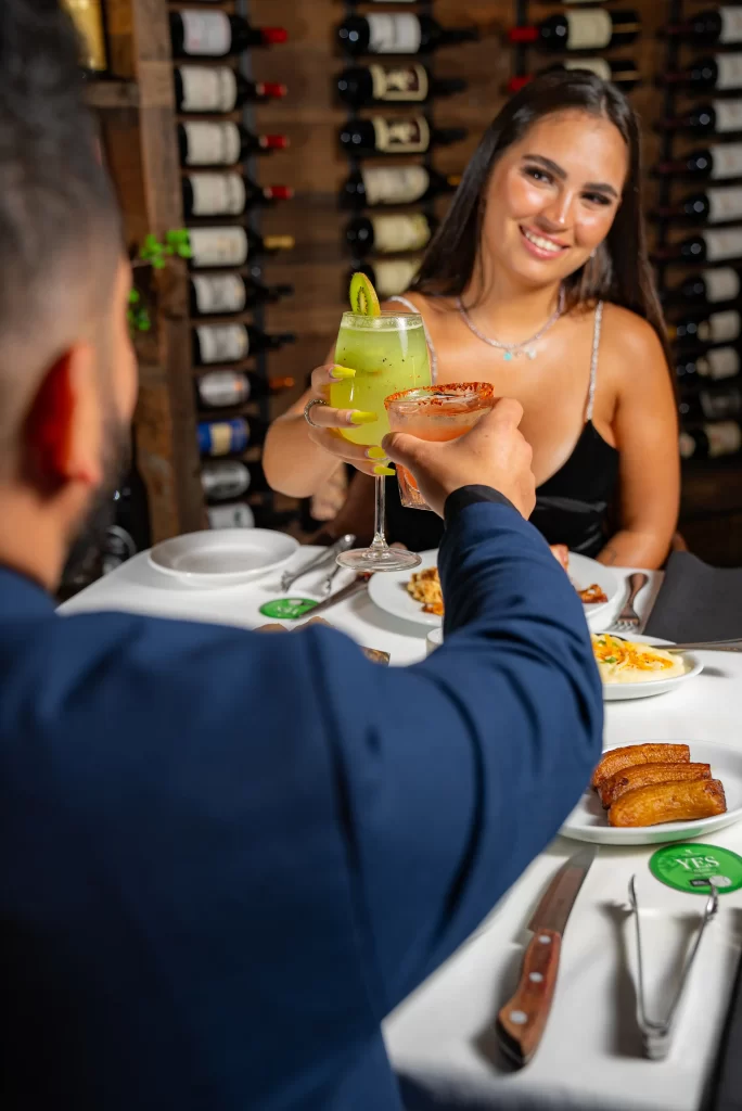 A woman in a black dress clinks glasses with a man, both smiling, against a wine-filled background, showcasing a celebratory moment.