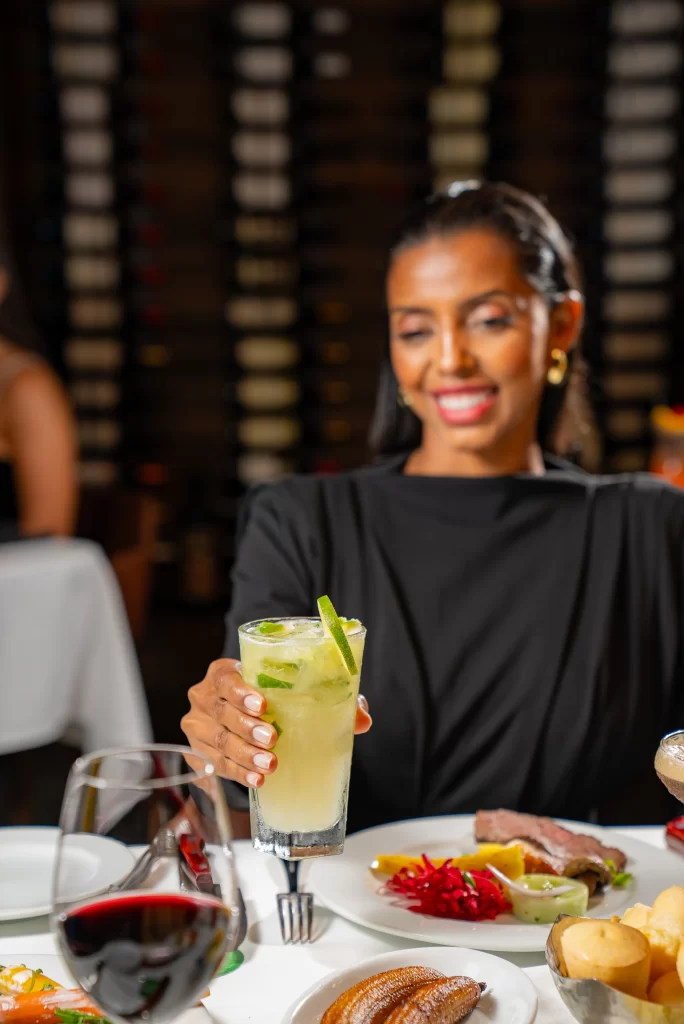 A woman holding a lime drink while seated at a table with a variety of dishes in a cozy dining atmosphere.