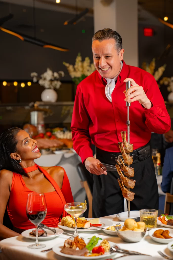 A server with a skewer of grilled meat interacts with a guest in an orange dress, who is smiling and enjoying her dining experience.