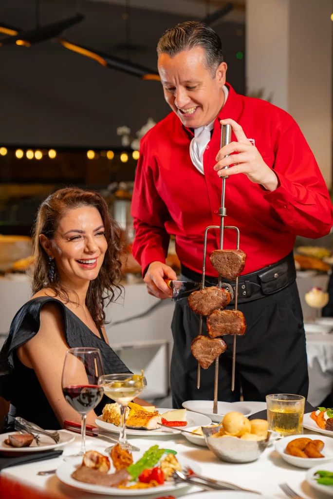 A server in red serving skewered meat to a smiling woman seated at a table, enjoying her meal with wine and side dishes.