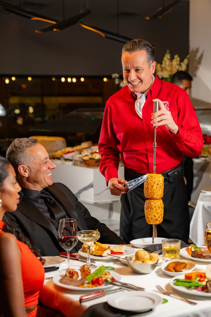 A server in a red shirt presents a skewer with roasted pineapple to two guests seated at a table filled with dishes and drinks, engaging in conversation.