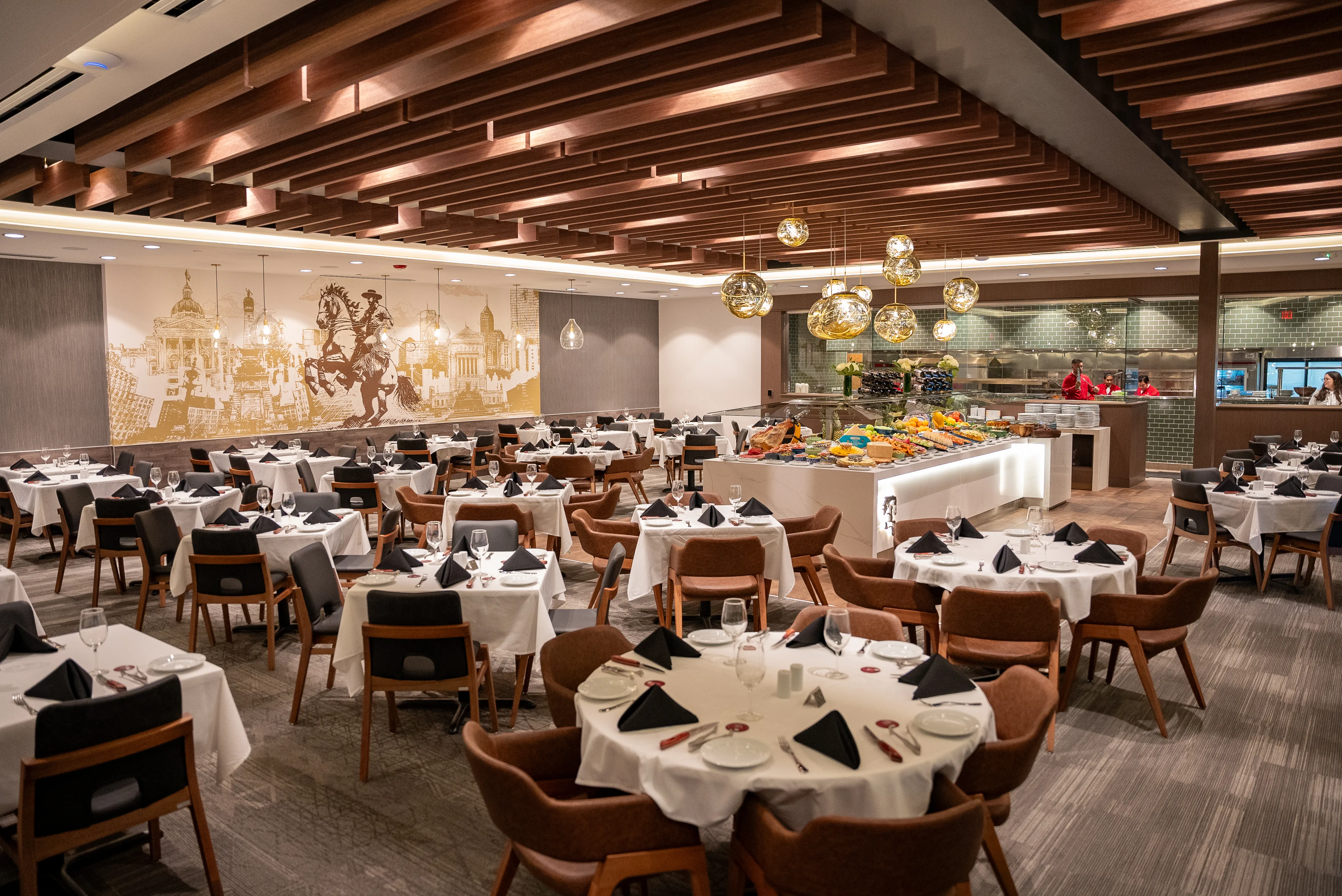 Upscale Brazilian steakhouse interior with white-clothed tables set with black napkins and wooden chairs, a central buffet island under decorative wood-beam ceiling accents, and an open kitchen in the background.
