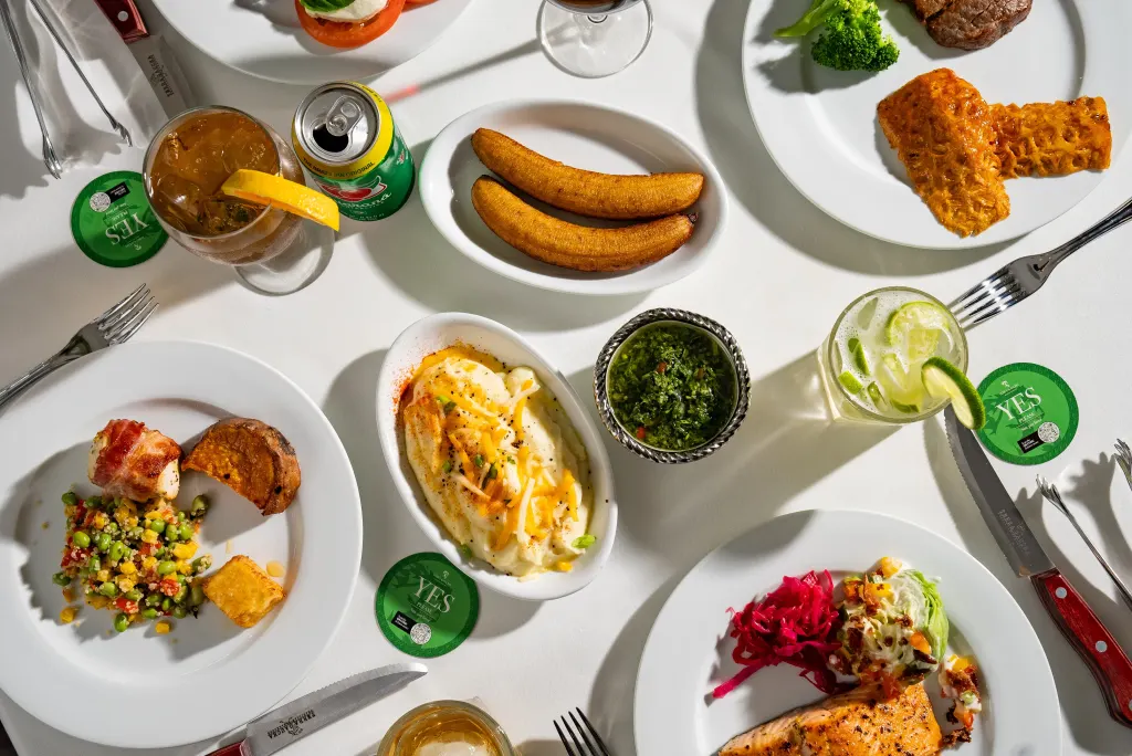 Overhead view of a restaurant table featuring grilled salmon, steak, fried plantains, mashed potatoes, fresh salads, vegetables, chimichurri, and cocktails.